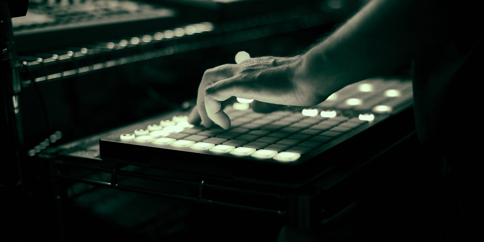 A hand pressing lit pads on a Launchpad in a dark room, with more gear on the shelves behind
